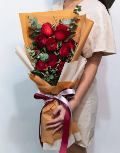 ผู้หญิงถือช่อดอกไม้ที่จัดด้วยดอกกุหลาบสีแดง 12 ดอก ห่อกระดาษสีส้มชานม ผูกริบบิ้นสีแดงและสีขาว