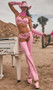 Sheriff Shine Cowgirl costume includes pink vinyl bra top with halter neck and back tie closure, mini booty shorts with puckered back, chaps, belt, bandana, and pair of long arm cuffs with fringe detail. Six piece set.