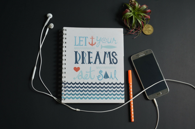 Journal with the title 'Let your dreams set sail' rests on a desk with mobile phone, headphones and pot plant