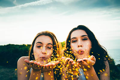 Two young women outdoors, blowing gold glitter from their hands