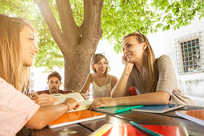 Group of students sit outdoors under a tree, with books spread out on a table