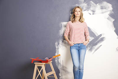 Young woman leans against a half-painted wall, with ladder and paintroller