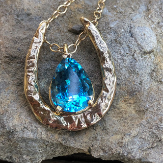 14k gold hammered horseshoe pendant. The shoe is elongated and reflects light playing on the piece. Gemstones are separate as are the chains. We can use your gemstone or one of ours. Pictured with topaz and green tourmaline. Call 8593330458 to order gemstones or chains. Gemstones and chains sold separately