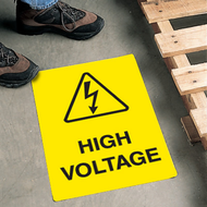 A picture of this black-on-yellow floor marker installed on a floor next to a person's feet and pallet. The sign has an ANSI high voltage symbol with wording "HIGH VOLTAGE" beneath the icon.