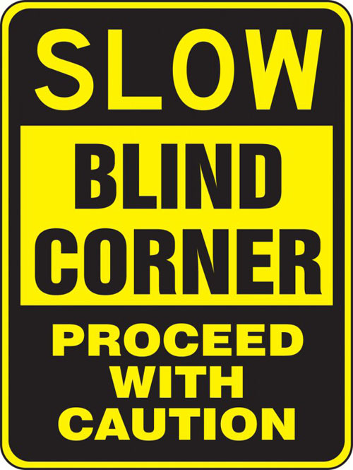 A photograph of a yellow and black 06254 blind corner sign, reading slow blind corner - proceed with caution.