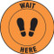 A photograph of an orange and black 11236 social distance floor sign, reading wait here with footprints icon.