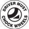 This black and white sign features the text "Driver Must Chock Wheels". The center shows the image of a chock being actively used on a wheel. Use to prevent accidents caused by uncontrolled vehicles.