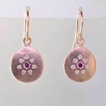 These sweet earrings shimmer when you wear them. Beautiful and warm 14 karat rose gold medallion drops sprinkled with .11ct. of ideal cut diamonds set in a starburst pattern, with a glowing pink sapphire in the center, hanging from wires. 12mm long.  

Designed and created in our studio by the artist Stuart J.