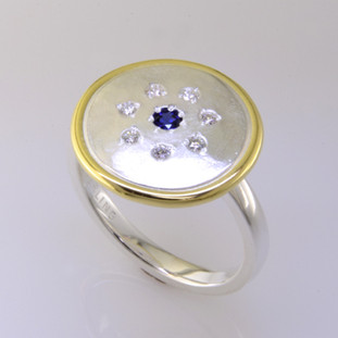 This sweet ring has a special, hand applied finish that shimmers when you wear it. Crafted in beautiful sterling silver, and framed in rich 18 karat yellow gold. It is sprinkled with 7 ideal cut diamonds weighing a tenth of a carat, with a Chatham® created deep blue sapphire gemstone in the center. This one is a size 7, but we can make it in any size. Ring top measures 5/8 inch in diameter.  

Chatham Gemstones are also available in Ruby, Sapphire, and Alexandrite.

Designed and created in our studio by the artist Stuart J.

Chatham® Created Gemstones are grown in carefully controlled laboratory conditions. They are chemically and optically identical to their natural counterparts, yet cost much less. Because the environment in which they are grown is controlled, the imperfections often found in natural gemstones, are absent in Chatham® Created Gemstones. Because of this, and superior cutting, Chatham® gems exhibit the color and sparkle of the finest natural gems. Each one comes with a Certificate of Authenticity, and a Lifetime Warranty.