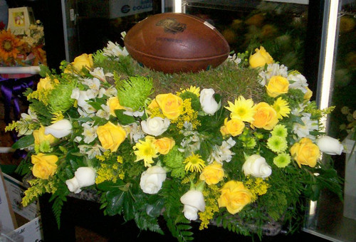 Football Player Casket Spray with football, yellow and white flowers with football placed on piece of grass. 